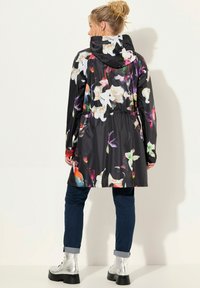 Zwart waterdicht regenjas met bloemenpatroon, capuchon, elastische taille en lange mouwen. Gecombineerd met donker denim met opgerolde mouwen en zilveren enkellaarzen.