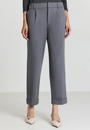 Femme portant un pantalon gris ajusté avec des ourlets retroussés et des talons aiguilles beiges, se tenant devant un fond clair.