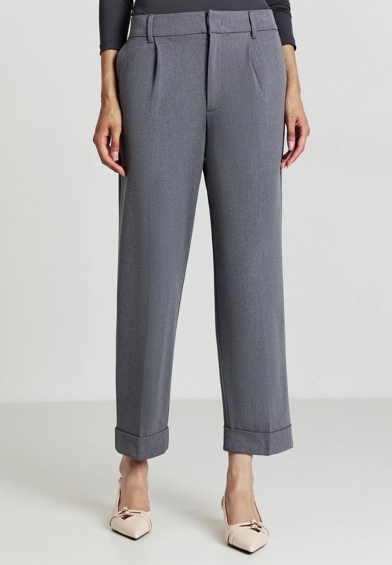 Donna che indossa pantaloni grigi su misura con orli risvoltati e décolleté beige a punta, in piedi davanti a uno sfondo chiaro e semplice.