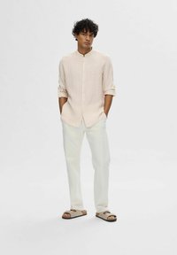 Camisa de lino de color rosa claro con cuello mao, pantalones blancos a medida y sandalias marrones. La camisa tiene botonería y mangas enrolladas.