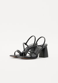 Sandales noires à talons hauts avec des fines lanières et un talon carré. Le design présente une finition lisse et une silhouette minimaliste.