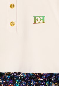 Polo shirt crema chiaro con due bottoni dorati e un logo "EH" multicolore; abbinato a una gonna nera decorata con motivi vivaci.