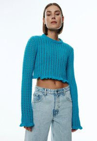 Blauer, gestrickter Crop-Pullover mit einer strukturierten, offenen Masche, langen Ärmeln und einem gewellten Saum. Kombiniert mit hochgeschnittenen, hellblauen Jeans.