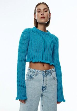 Blauer, gestrickter Crop-Pullover mit einer strukturierten, offenen Masche, langen Ärmeln und einem gewellten Saum. Kombiniert mit hochgeschnittenen, hellblauen Jeans.