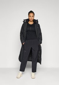 Woman standing with hands in pockets wearing a long black puffer coat, black pants, black shirt, white socks, and beige sneakers on a gray background.