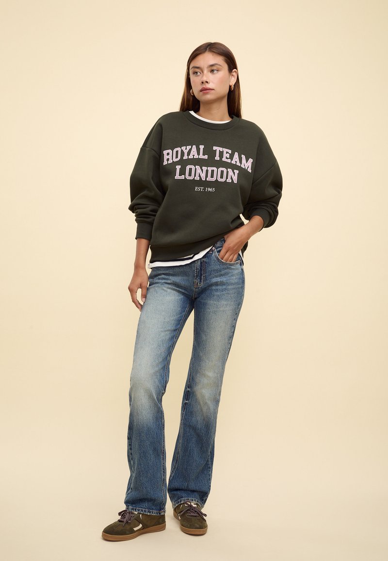 Giovane donna con felpa verde scuro "Royal Team London", jeans blu e scarpe da ginnastica verde oliva, in piedi con la mano in tasca davanti a uno sfondo beige.