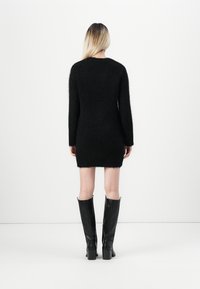 Kvinne med langt blondt hår iført en svart, lodden minikjole med lange ermer og svarte skinnstøvler som går til knærne, stående med ryggen mot.