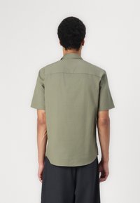 Chemise olive verte à manches courtes, avec un col standard, une texture lisse et un ourlet arrondi. Présente des coutures simples sans accents supplémentaires.