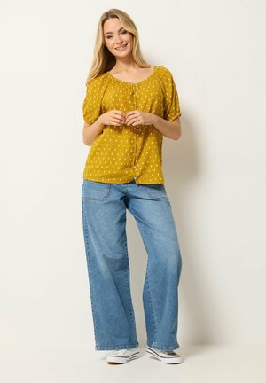 Uśmiechnięta kobieta stoi, mając na sobie musztardowo-żółtą bluzkę w wzory, szerokie niebieskie dżinsy i białe trampki na jednolitym jasnym tle.