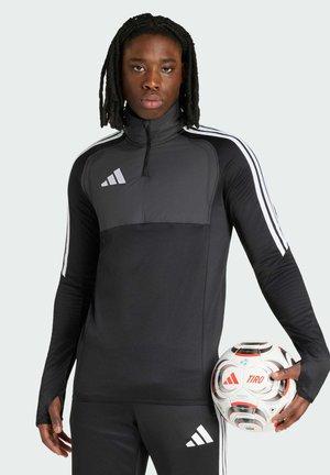 Junger Mann mit Dreadlocks trägt schwarzen Adidas Trainingsanzug und hält einen weißen Adidas Tiro Fußball mit roten Akzenten.