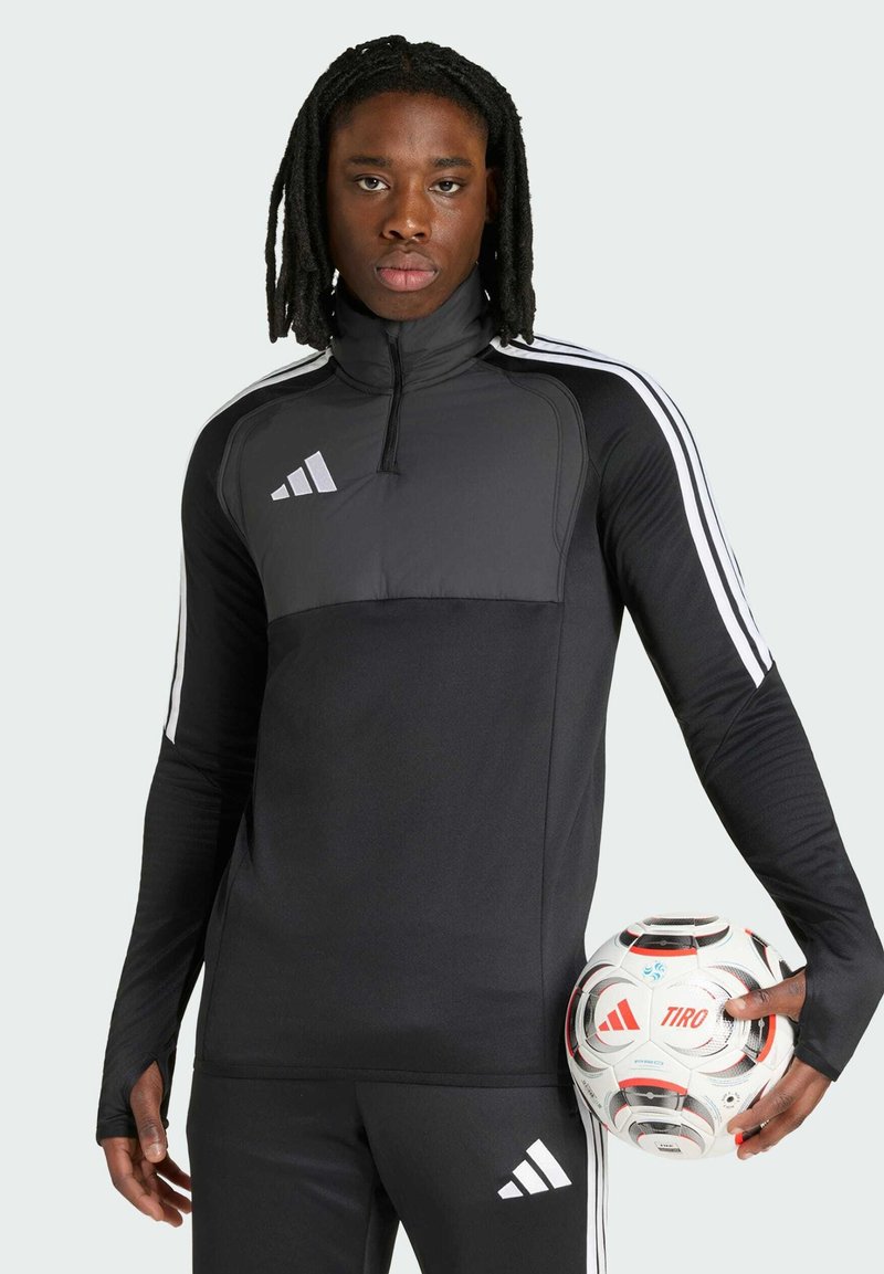 Jeune homme avec des dreadlocks portant un survêtement noir Adidas tenant un ballon de football Adidas Tiro blanc avec des accents rouges.
