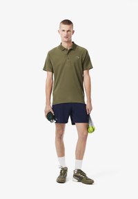 Oliivinvihreä polo-paita lyhyillä hihoilla, laivastonsiniset shortsit ja oliivinvihreät tennarit. Pitelee vihreää pyyhettä ja tennispalloja verkossa olevassa laukussa.