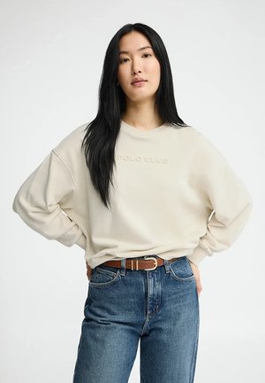 Aziatische vrouw met lang zwart haar, gekleed in een beige sweatshirt en een blauwe spijkerbroek met een bruine riem, staand met de handen op de heupen tegen een witte achtergrond.