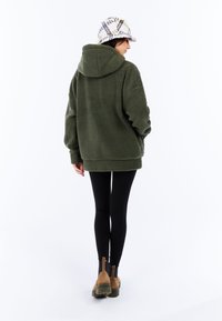 Haut à capuche en tricot olive surdimensionné avec une finition texturée, doté d'une capuche, de manches longues et de fentes sur les côtés à l'ourlet. Porté avec des leggings noirs.