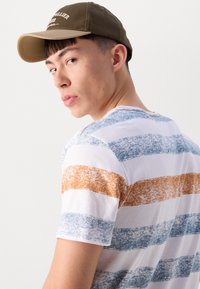 Camiseta a rayas con rayas texturizadas en blanco, azul y naranja; cuello redondo y mangas cortas. Se lleva con una gorra marrón.