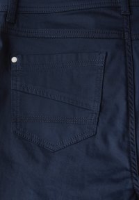 Námořnické modré kalhoty s zadní kapsou ve tvaru diamantu, viditelným švem a malým bílým knoflíkem v horním rohu.