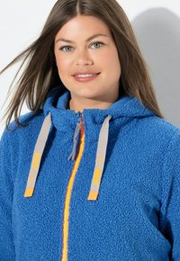 Blauer Fleece-Hoodie mit einer weichen, strukturierten Oberfläche. Verfügt über einen orangefarbenen Reißverschluss, graue Kordeln mit Logo und eine Kapuze.