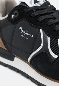 Μαύρο αθλητικό παπούτσι από σουέτ και νάιλον υλικά. Περιλαμβάνει λευκές λεπτομέρειες, υφές με μοτίβα και ένα λογότυπο τυπωμένο στο πλάι.