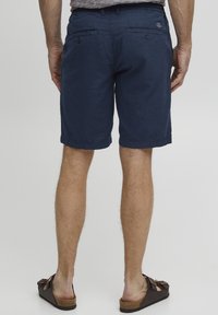 Mørkeblå shorts med løs passform, med to baklommer med knapper og en ensfarget tekstur. Brukt med brune tøfler.