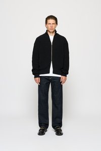 Zwarte, geribbelde gebreide zip-up cardigan met een hoge kraag, gedragen over een wit t-shirt, gecombineerd met donkere jeans en zwarte instappers.