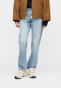 Φωτεινό μπλε τζιν με ίσια γραμμή, φορεμένο με κρεμ αθλητικά παπούτσια και καφέ ζακέτα με φερμουάρ πάνω από μαύρο τοπ.