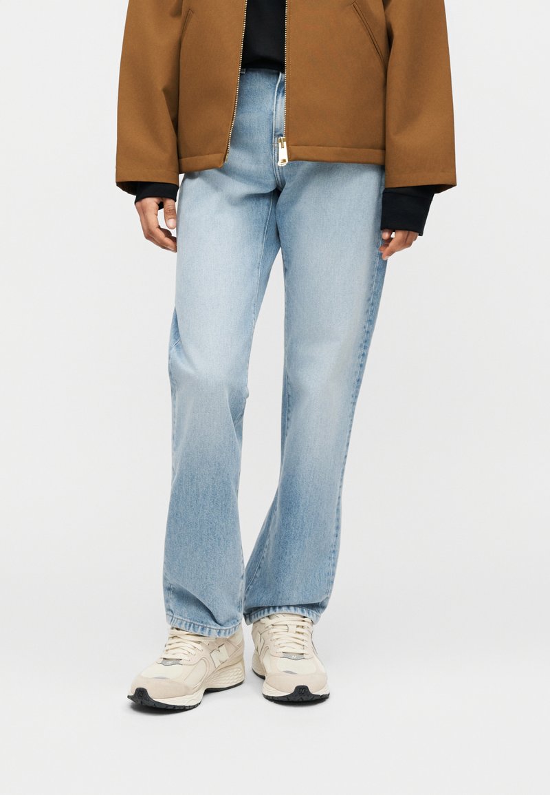 Lichtblauwe jeans met rechte pijpen, gedragen met crèmekleurige sneakers en een bruine jas met rits over een zwart shirt.