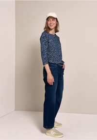 Blaue gemusterte Langarmbluse, dunkle Jeans und helle Sneaker. Modell trägt eine weiße Kappe und steht vor einem neutralen Hintergrund.