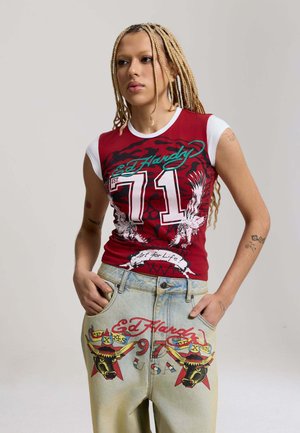 Persoon met gevlochten haar, gekleed in een rood-wit Ed Hardy-shirt en lichte jeans met kleurrijke grafische prints, handen in de zakken, neutrale achtergrond.