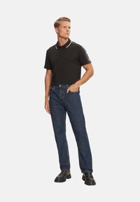 Camisa de polo negra con detalles en el cuello blanco, combinada con jeans rectos azules y zapatos negros. El modelo está de pie con las manos en los bolsillos.