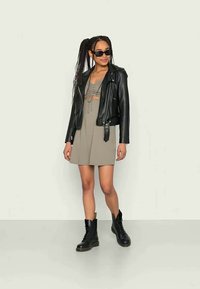 Mulher com tranças longas usa óculos de sol pretos, jaqueta de couro preta, vestido taupe e botas pretas com cadarços, de pé em frente a um fundo simples.