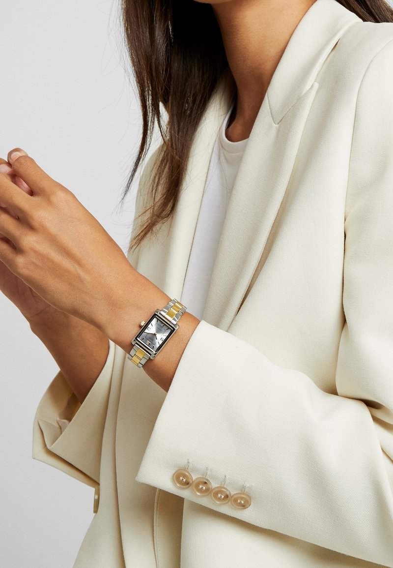 Femme en blazer blanc portant une montre-bracelet rectangulaire bicolore argent et or à son poignet gauche sur un fond uni.
