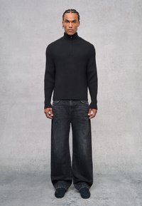 Schwarzer gerippter Pullover mit hohem Kragen und Viertelreißverschluss. Getragen mit locker sitzenden dunklen Jeans und dunklen Schuhen. Hintergrund ist strukturiertes Grau.