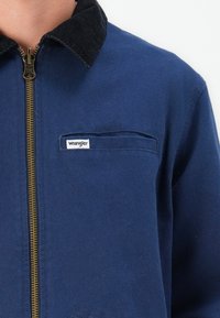 Chaqueta con cremallera de color azul marino, hecha de tela duradera, que presenta un cuello de pana, un bolsillo en el pecho y un parche de logo visible en la parte superior izquierda.