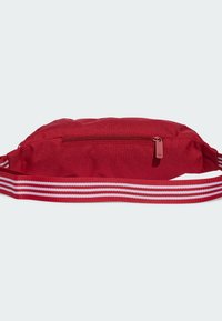 Červená látková ledvinka s kapsou na zip, vybavená nastavitelným popruhem se červenými a bílými pruhy. Hladká textura, kompaktní design.