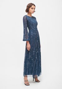Robe longue bleu marine en tissu transparent, ornée de motifs de perles complexes, avec des manches évasées et une taille cintrée accompagnée d'une jupe fluide.