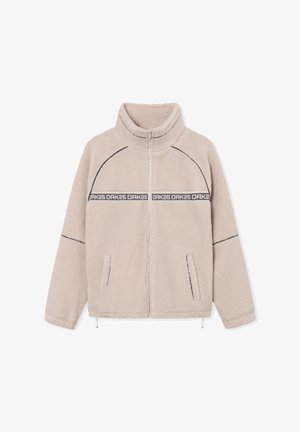 Beige Fleecejacke mit hohem Kragen, Reißverschluss, Seitentaschen und kontrastierender schwarzer Naht. Mit einem Logo-Band über der Brust.