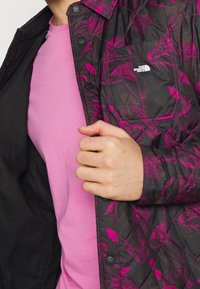 Quiltad jacka med mörk bas och rosa blommönster, med en ficka och knappframsidan, bärs över en enfärgad rosa skjorta.