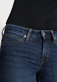 Jeans de mezclilla azul oscuro con corte ajustado, con costuras amarillas, bolsillos frontales y cierre de botón metálico en la cinturilla.