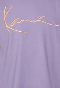 Levandulové tričko s hladkou texturou a vyšívaným kurzívním logem v kontrastní oranžové barvě umístěným uprostřed na přední straně.