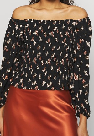 Vrouw draagt een zwarte off-shoulder bloemenblouse met gerimpeld lijfje en lange mouwen, gecombineerd met een roestkleurige satijnen rok.