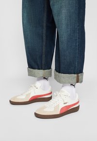 Bílé a béžové tenisky s červeným pruhováním, vyrobené z jemné kůže a semiše, s kulatou špičkou, hnědou gumovou podrážkou, nošeny s ohrnutými modrými džíny.