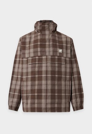 Jeune homme portant une veste à capuche à carreaux marron et blanc avec une grande poche devant et un pantalon noir sur fond blanc uni.