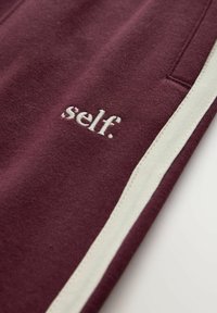 Bordeaux broek met witte zijstrepen en geborduurd logo "self." nabij de zak. Zacht materiaal met een gladde textuur.