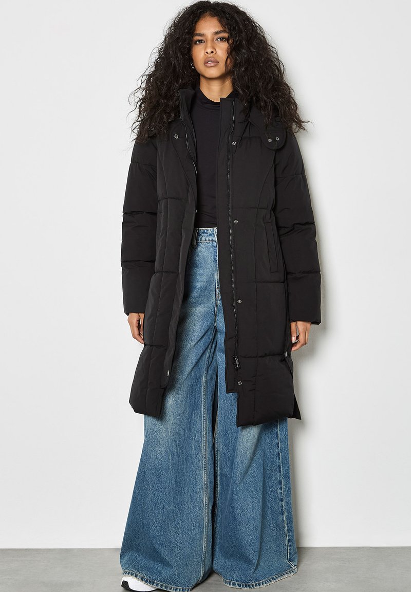 Cappotto imbottito nero con chiusura a zip e automatico, dotato di collo a imbuto e tasche laterali. Abbinato a jeans blu a gamba larga.