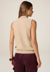 Suéter sin mangas de punto beige con cuello y dobladillo acanalados, combinado con pantalones de pana burdeos oscuros. Textura suave con un brillo sutil.