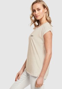 Beige t-skjorte med korte ermer laget av bomull og rund hals. Har teksten "Inspire" i svart på forsiden, med en avslappet passform.