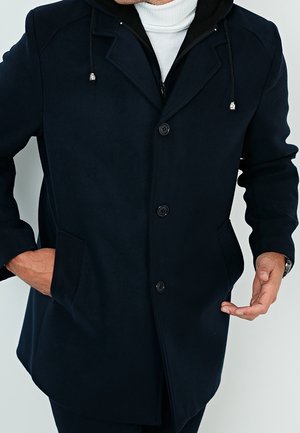 Mies pukeutunut tummansiniseen takkiin, jossa napit ja sivutaskut, valkoisen pooloneuleen päällä, toinen käsi taskussa ja toinen elehtii.