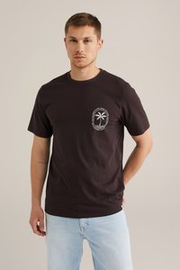 Zwarte katoen T-shirt met een palmboomafbeelding op de linker borst. Ronde hals, korte mouwen, ontspannen pasvorm. Lichtblauwe denim jeans.