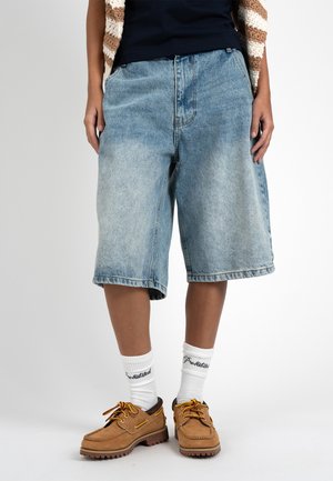 Person trägt blaue Denim-Longshorts, weiße Socken mit schwarzem Text, braune Schnürschuhe und ein schwarzes Top mit einem braun-weiß gestreiften ärmellosen Cardigan.