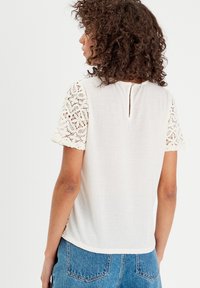 Haut blanc à manches courtes avec des manches en dentelle délicate et un détail ajouré au dos. Texture lisse, coupe décontractée et ourlet légèrement arrondi.